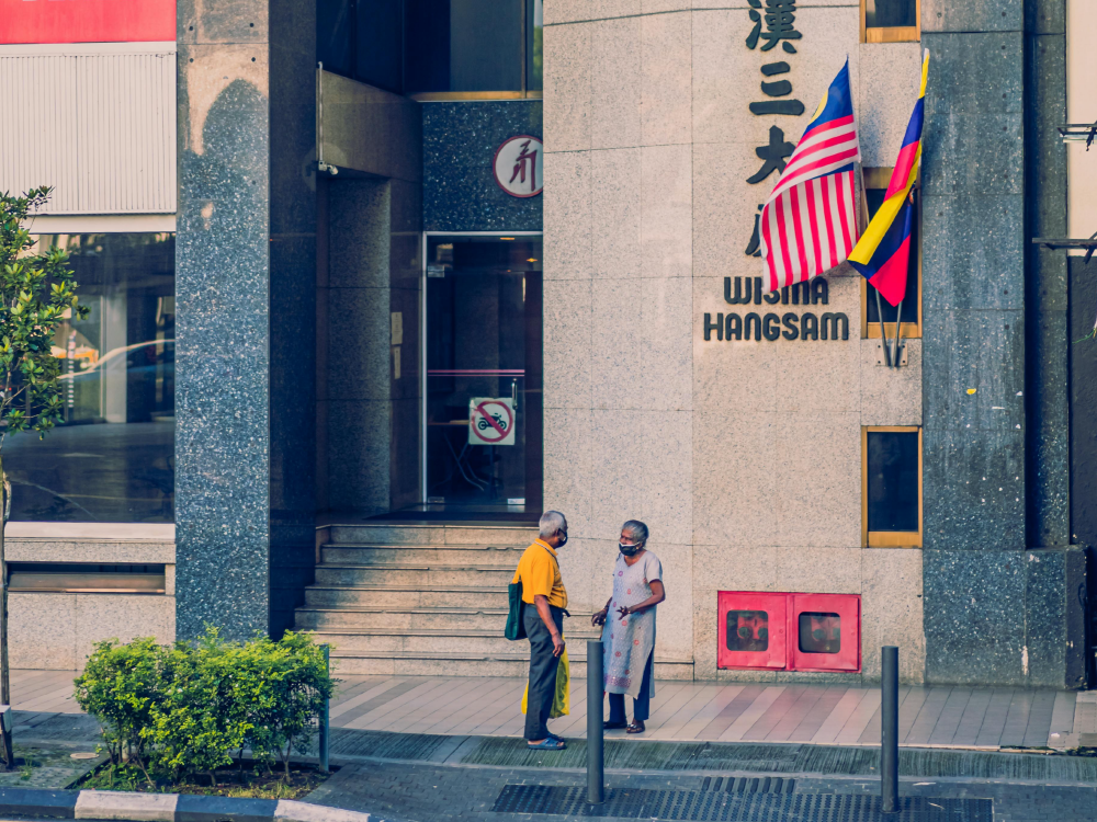 Two people in masks converse outside a building with flags